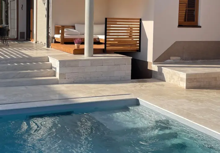 Piscina con bordo in pietra travertine. Gradini e pavimentazione in travertine creano un'elegante area relax con divani in legno.