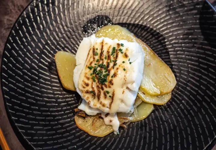 Bacalao gratinado con alioli, servido sobre patatas doradas en un plato negro texturizado.