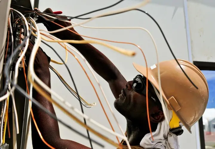 Elektryk w kasku i okularach pracuje przy skrzynce z przewodami. Nauka jak zostać elektrykiem wymaga precyzji.