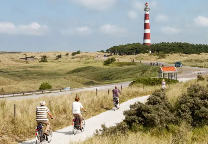 Ciclistas disfrutan de un paseo por la ruta EuroVelo 7, con un faro a rayas rojas y blancas al fondo.
