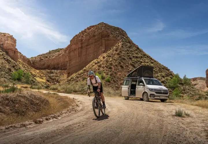 Ciclista pedaleando por un camino de badlands gravel, con una furgoneta camper aparcada al fondo.