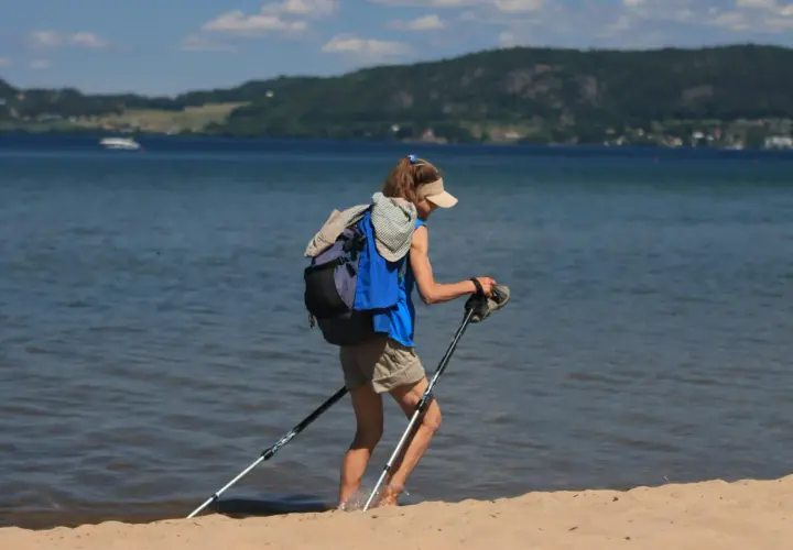 Osoba z plecakiem i kijkami do nordic walking nad morzem, wchodząca do wody.