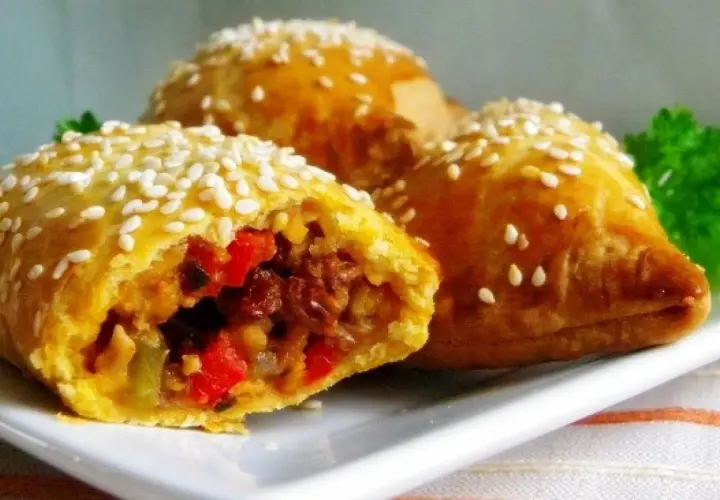 Złociste empanadas posypane sezamem, z widocznym farszem z warzyw i mięsa. Pyszna przekąska.
