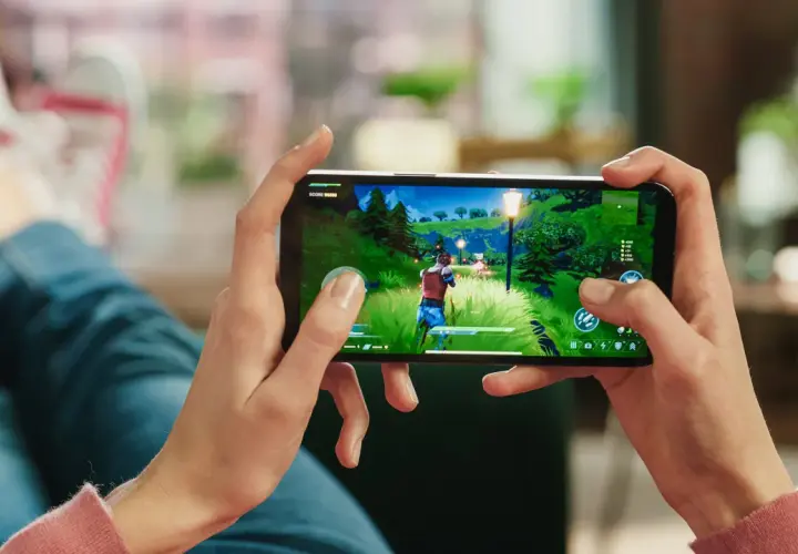 Ręce trzymają smartfon, na którym widać fragment popularnej gry na telefon. W tle widać nogi w jeansach i trampkach.