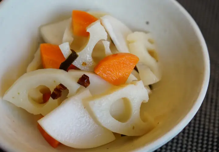 A close-up of a bowl filled with a colorful mix of Japanese root vegetables, including sliced lotus root with its distinctive holes, bright orange carrot pieces, and possibly daikon radish.