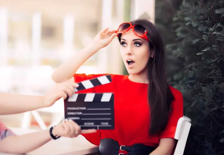 Aktorka w czerwonej sukience z okularami na głowie, zszokowana, gdy klaps filmowy zbliża się do jej twarzy. Stopniowanie high.