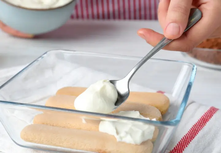 Nakładanie łyżką puszystego kremu z mleka skondensowanego i mascarpone na biszkopty w szklanej formie.