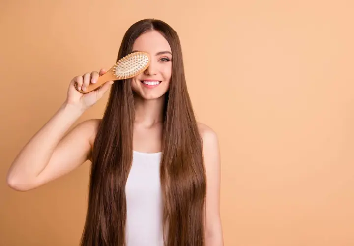 Mujer sonriente con pelo largo y liso, sosteniendo un cepillo de madera sobre su ojo. ¡Un truco para saber como desenredar un pelo muy enredado!