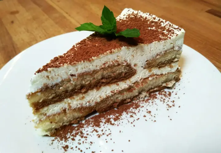 Pyszny kawałek najprostszego tiramisu, posypany kakao i udekorowany listkiem mięty, na białym talerzu.