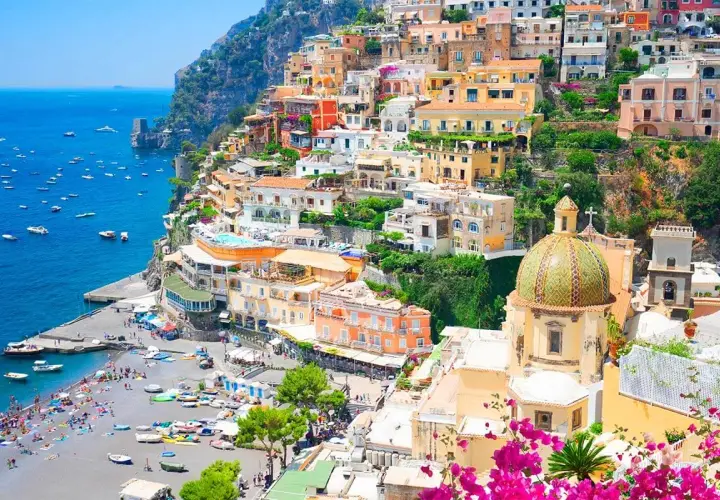 Colorful towns near Naples, Italy, cascade down a cliff to a busy beach and blue sea. A domed church stands out.