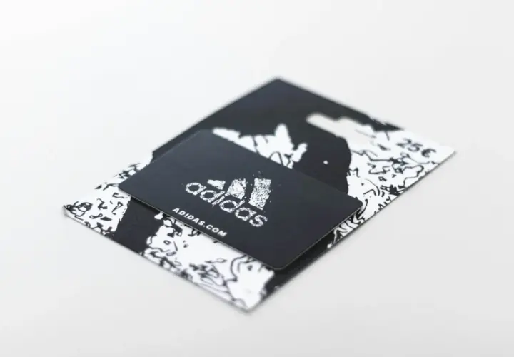 Czarny voucher Adidas z logo i adresem strony internetowej. Dowiedz się, jak zrobić voucher na prezent.