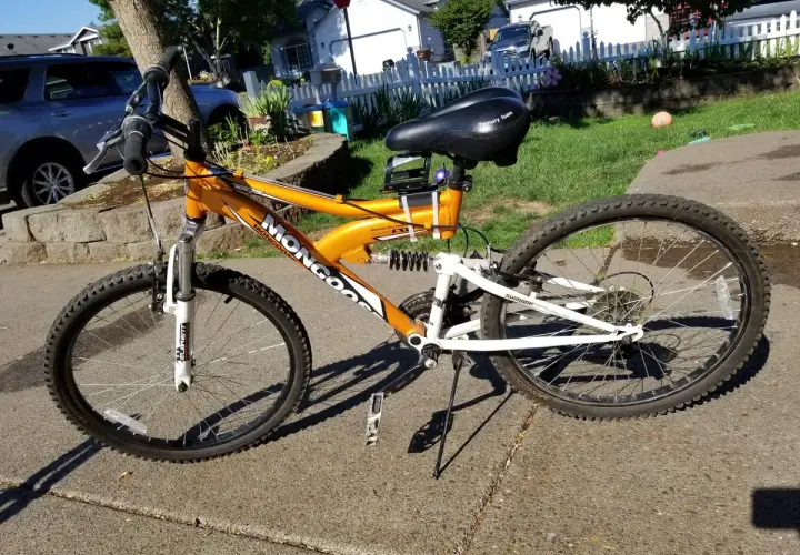 An orange Mongoose XR-75 mountain bike with a black seat and white suspension sits on a sidewalk.