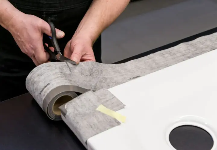Hände schneiden Dichtungsband zu, um eine Duschwanne abzudichten. Das Material ist auf einer Rolle und wird präzise zugeschnitten.