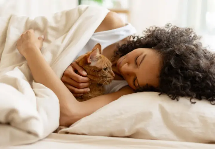 Frau mit Katze kuschelt im Bett unter einer Decke.