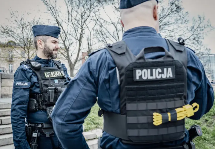 Dwóch policjantów w mundurach i kamizelkach taktycznych. Widoczne wyposażenie policjanta: radiostacja, kabura na broń, pas z ładownicami.