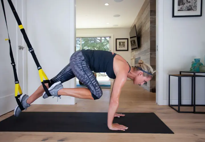 Kobieta wykonuje ćwiczenie z taśmami TRX, angażując mięśnie brzucha i nóg. Intensywny trening TRX w domowym zaciszu.