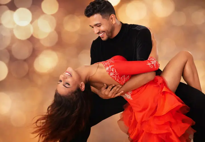 Coppia che balla con passione una salsa ballo latino, lei in abito rosso, lui in nero, su sfondo sfocato con luci dorate.