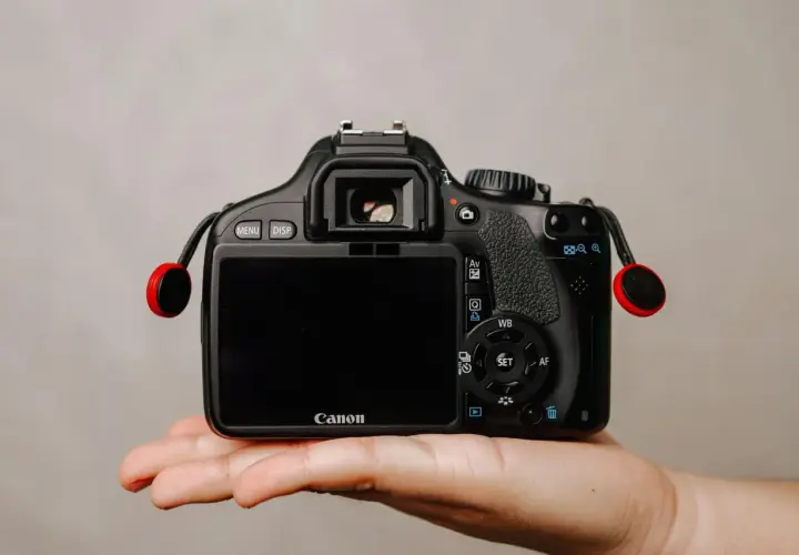 Dłoń trzyma czarną lustrzankę Canon z czerwonymi elementami smyczy.