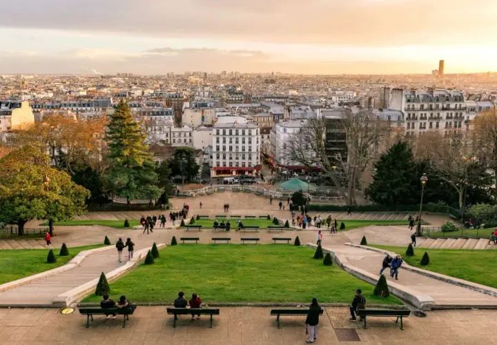 Widok z wzgórza Montmartre na Paryż. Ludzie spacerują po zielonych tarasach i odpoczywają na ławkach, podziwiając panoramę miasta.