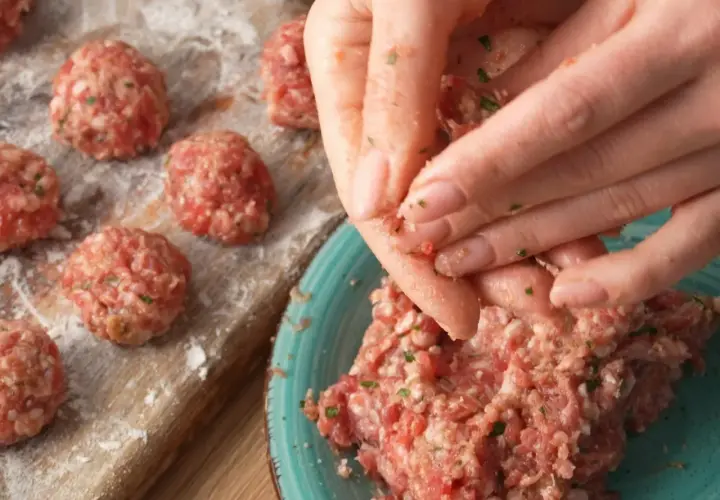 Ręce formują klopsiki według przepisu na meatballs. Na desce widać już gotowe, obtoczone w mące kulki mięsa.