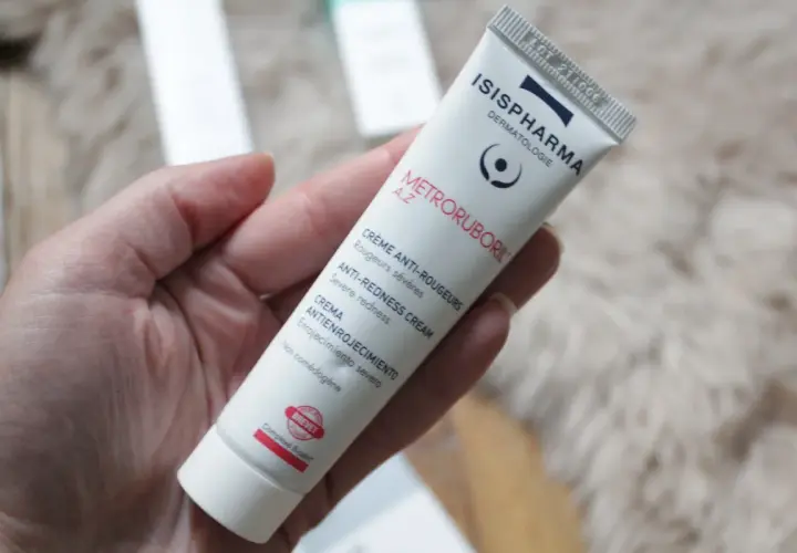 Une main tient un tube de crème anti-rougeurs de la marque Isispharma, contenant de l'acide azélaïque pour apaiser les rougeurs sévères.