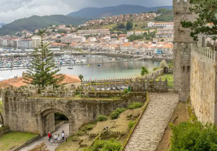 View from a castle wall overlooking a harbor filled with boats, a historic ship, and a town. The path leads through an ancient stone archway, hinting at a journey from porto to Baiona.