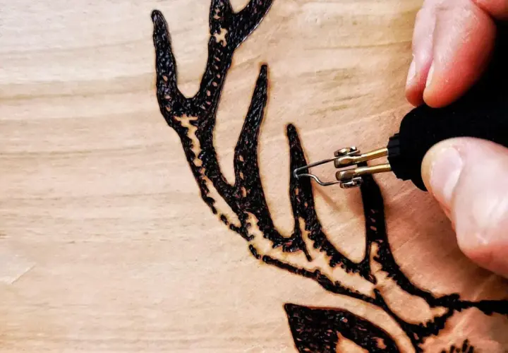 Dettaglio di lavori con pirografo su legno: una mano che disegna un palco di cervo con precisione.