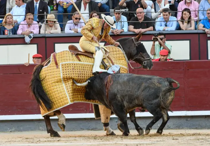 Un torero a caballo, con traje de luces, se enfrenta a un toro en la arena. El caballo lleva una manta amarilla. El público observa desde las gradas.