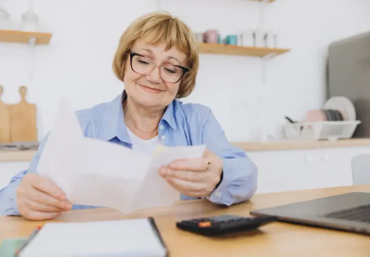 Uśmiechnięta starsza pani analizuje dokumenty, być może dotyczące ponownego przeliczenia emerytury ZUS. Obok leży kalkulator i laptop.