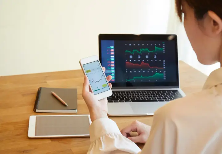 Frau analysiert Aktienkurse auf Laptop und Smartphone. Sie plant, wie sie ihre Aktiengewinne versteuern kann, während sie Notizen macht.