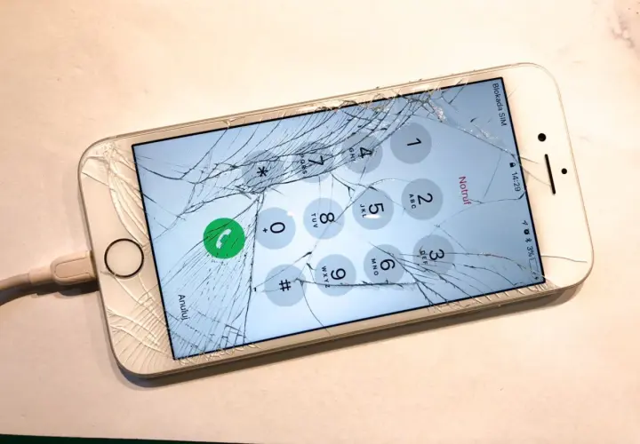 Zepsuty ekran telefonu z widocznym pęknięciem, podłączony do ładowarki. Na ekranie widoczna klawiatura numeryczna.