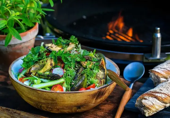 En stor skål med god sallad, grillad aubergine och sparris, med en grill i bakgrunden. Perfekt för en sommarkväll.