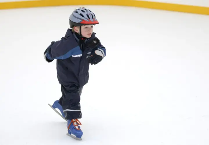 Bambino con casco e tuta blu pratica il pattinaggio sul ghiaccio, con i pattini azzurri e lacci arancioni.