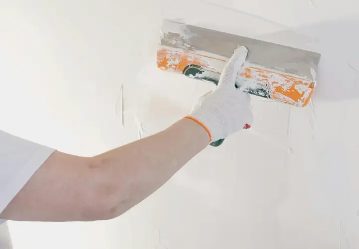 Mano con guanto bianco stende stucco murale su una parete bianca con una spatola arancione.
