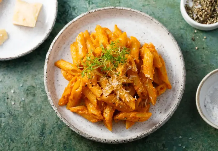 Penne z aromatycznym, czerwonym pesto, posypane parmezanem i świeżymi ziołami. Obok miseczka z solą i pieprzem.