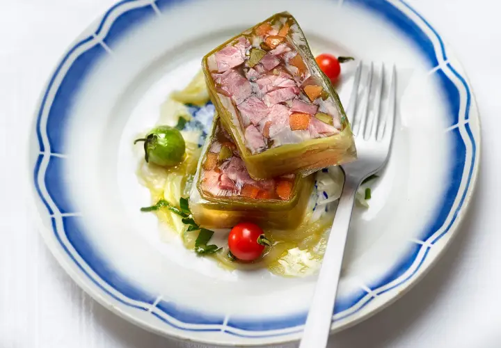 Deux portions de fromage de tête translucide, garnies de légumes et de viande, sur une assiette ornée de bleu.