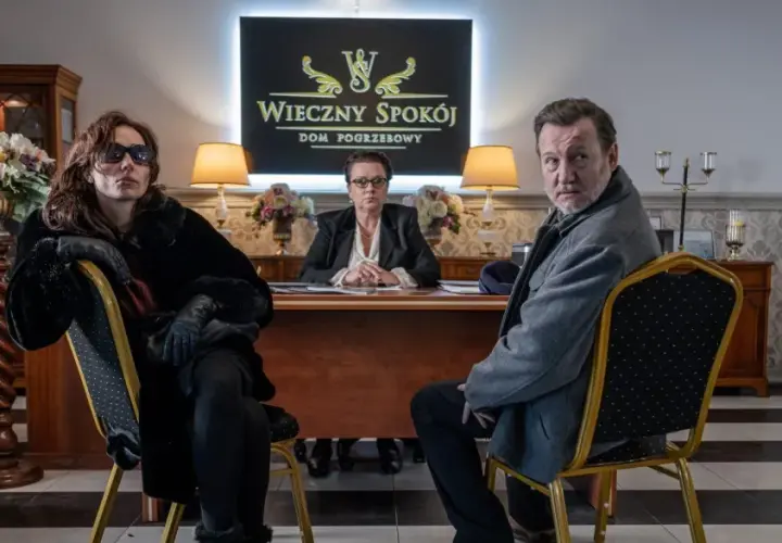 Trzy osoby siedzą w eleganckim biurze domu pogrzebowego. W tle logo "Wieczny Spokój". Scena z serialu "Minuta ciszy sezon 2".