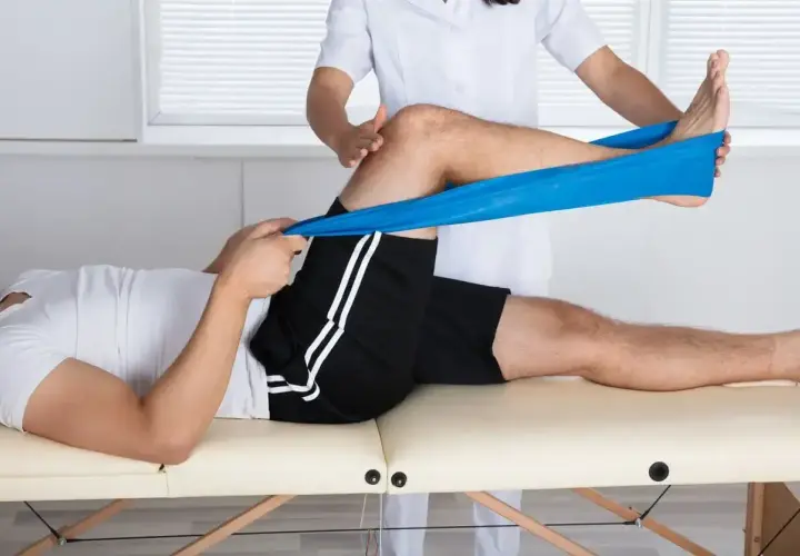 Fizjoterapeuta pomaga pacjentowi w ćwiczeniach mięśnia czworogłowego uda z użyciem taśmy oporowej.
