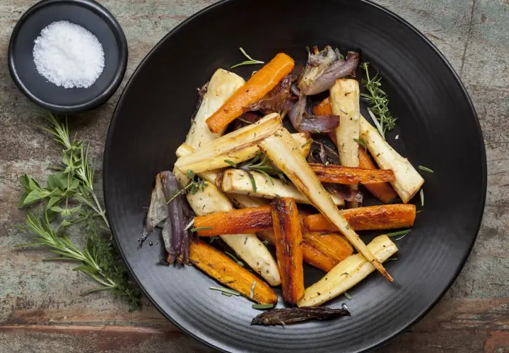 Légumes rôtis, carottes et panais, parsemés d'herbes. Un navet doré, tendre et savoureux, se mêle aux autres légumes sur l'assiette noire.