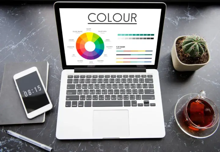 Na ekranie laptopa widać koło barw i palety kolorów, prezentujące różnice między cmyk vs rgb. Obok telefon, notes i filiżanka herbaty.