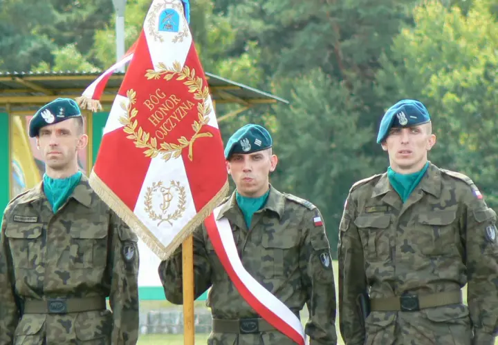 Trzech żołnierzy rezerwy w mundurach i beretach stoi obok sztandaru.