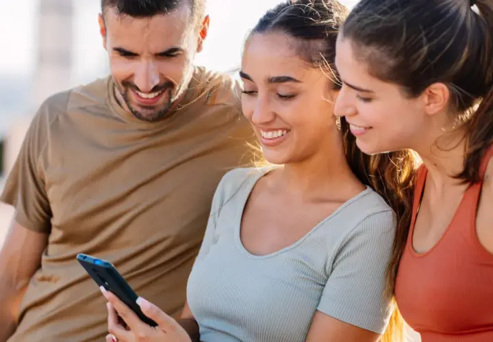Trzech przyjaciół ogląda coś na telefonie. Uśmiechnięta dziewczyna, która trzyma telefon, może ukrywać zakochanie, gdy patrzy na ekran.