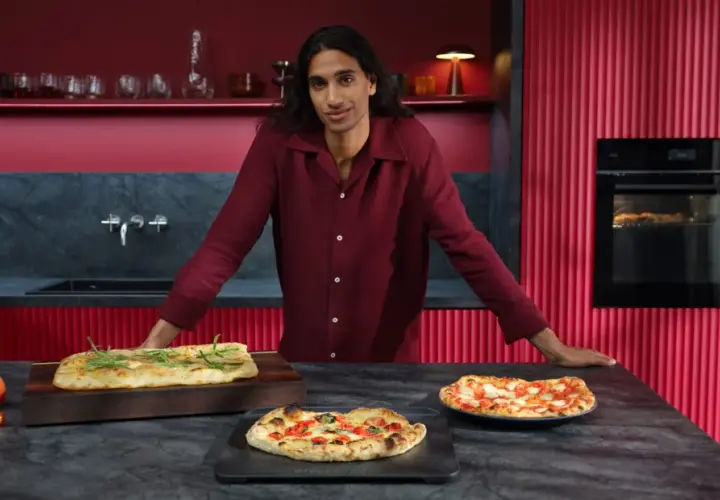 Mann in bordeauxrotem Hemd präsentiert frisch gebackene Pizzen. Im Hintergrund ein Ofen mit Pizzafunktion, der eine weitere Pizza bäckt.