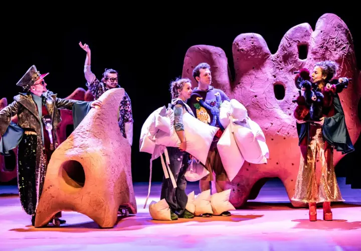 Aktorzy w barwnych strojach, niosący poduszki, na tle scenografii przypominającej gigantyczne, różowe formacje. To fragment spektaklu z repertuaru teatru lalki.