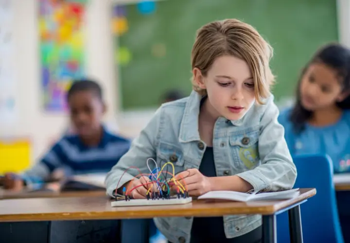 Ein Mädchen baut in einem Klassenzimmer an einem Stromkreis. Dies ist ein Beispiel für kompetenzorientierten Unterricht, bei dem Schüler praktische Fähigkeiten erlernen.