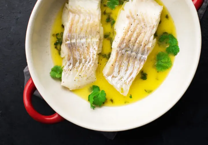 Dwa filety halibut pieczony ze skórą w maśle z ziołami i kolendrą, podane w białej misie z czerwonymi uchwytami.