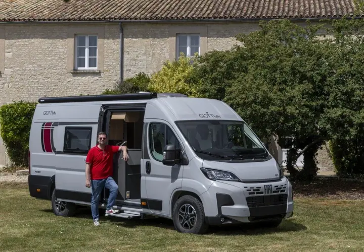 Un homme pose devant un **fourgon ou camping car** gris, porte ouverte, devant une maison en pierre et des arbres.