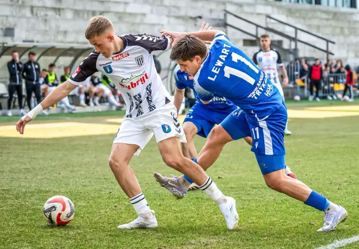 Piłkarze walczą o piłkę na boisku. Jeden w białej koszulce, drugi w niebieskiej z numerem 11. To ważny mecz, który może wpłynąć na 3. liga tabelę.