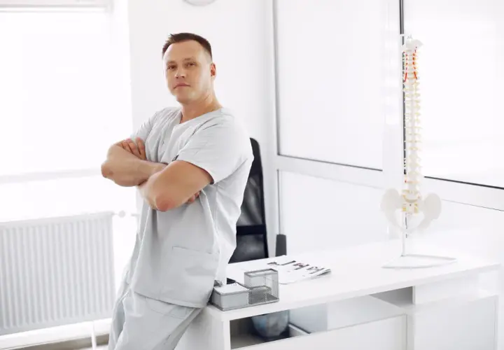 Mężczyzna w uniformie medycznym opiera się o biurko, na którym stoi model kręgosłupa. To przyszły specjalista, który wybrał fizjoterapia studia.