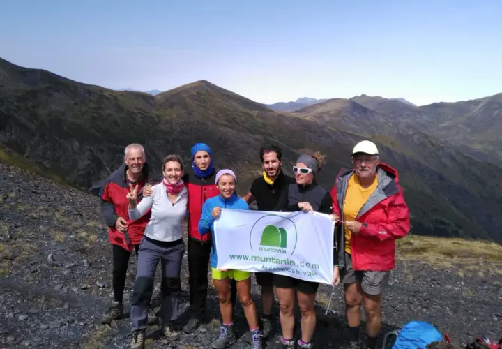 Grupo de senderistas celebra en la cima de una montaña, con el valle de **Aran** de fondo. Sostienen una pancarta de Muntania.com.